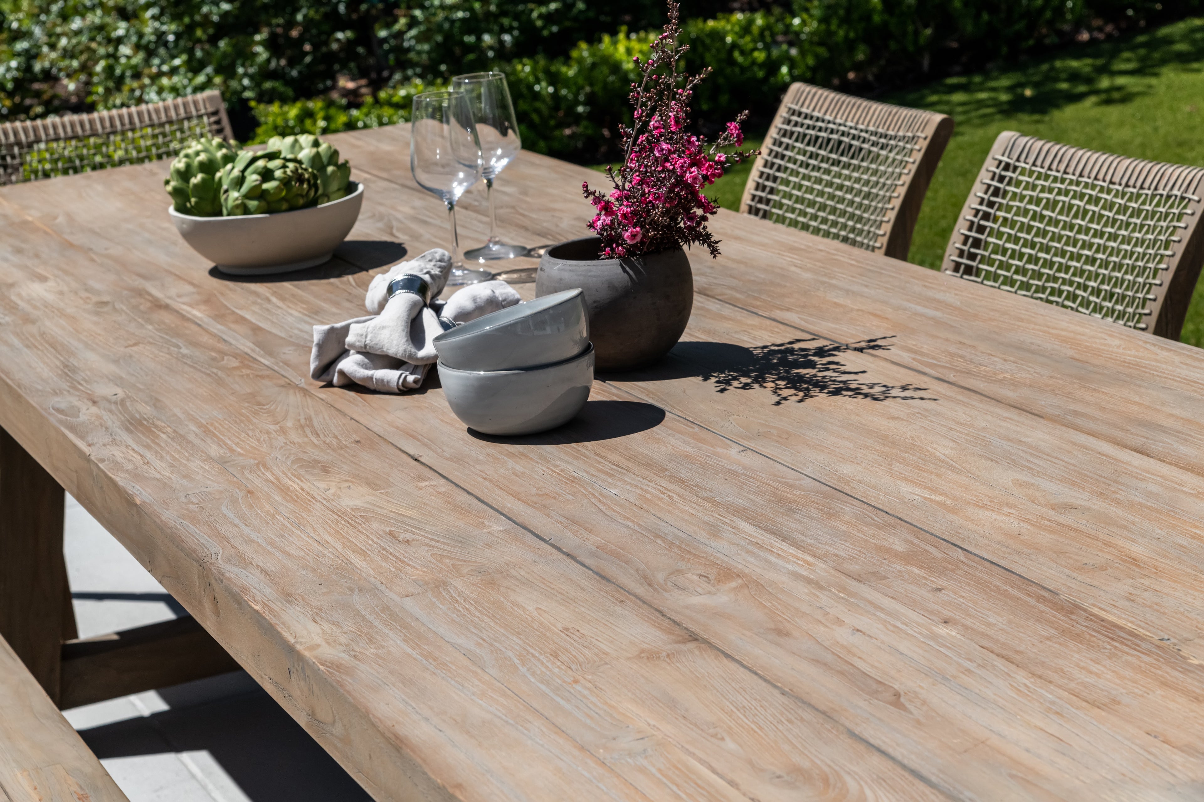 A wooden table is set outdoors with stacked bowls, a vase of pink flowers, artichokes in a bowl, linen napkins, and wine glasses. It’s surrounded by wicker chairs in a garden setting.