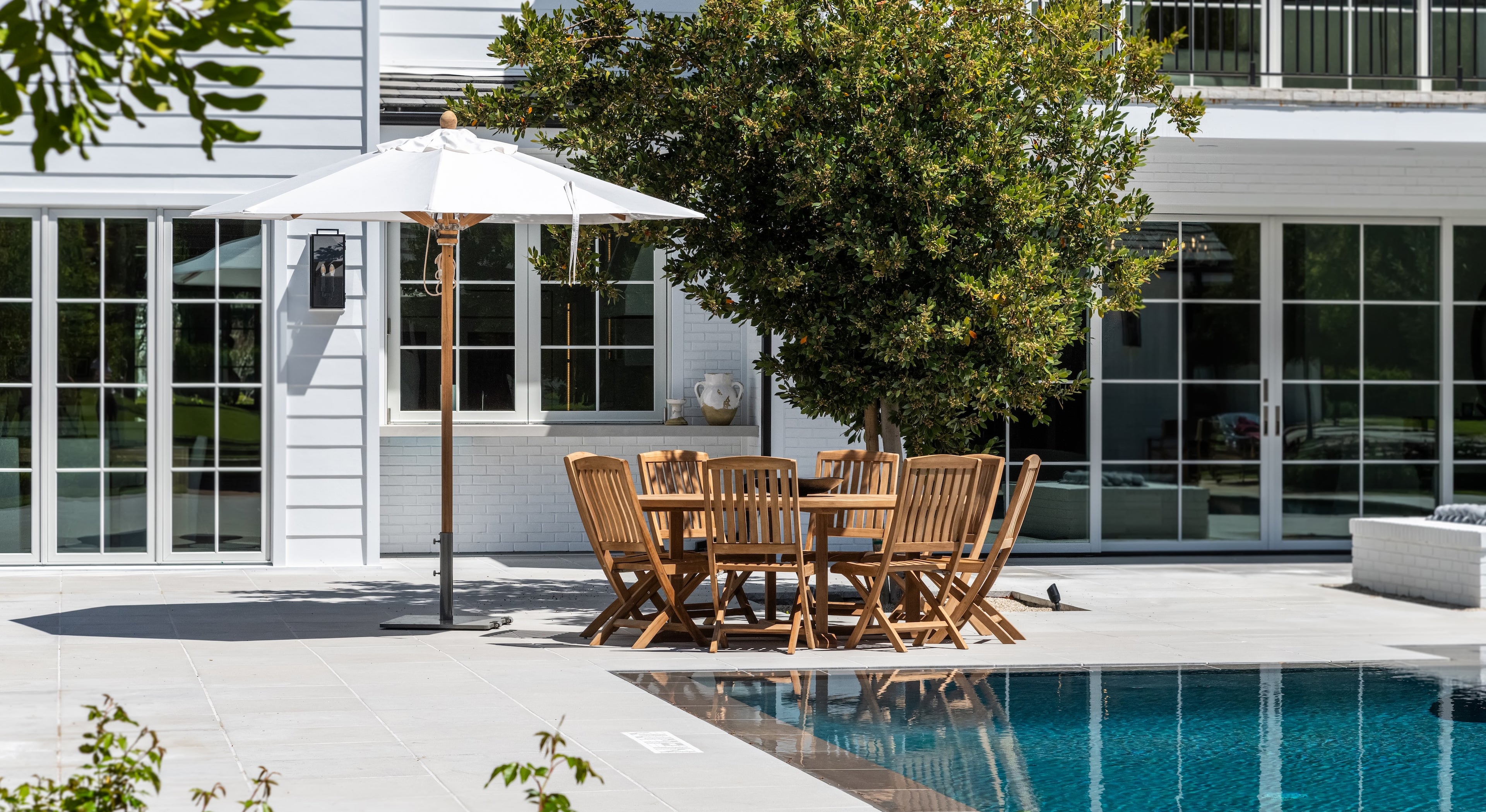 A wooden dining table with chairs is set under a white umbrella beside a swimming pool, adjacent to a white house with large windows and a lush tree.