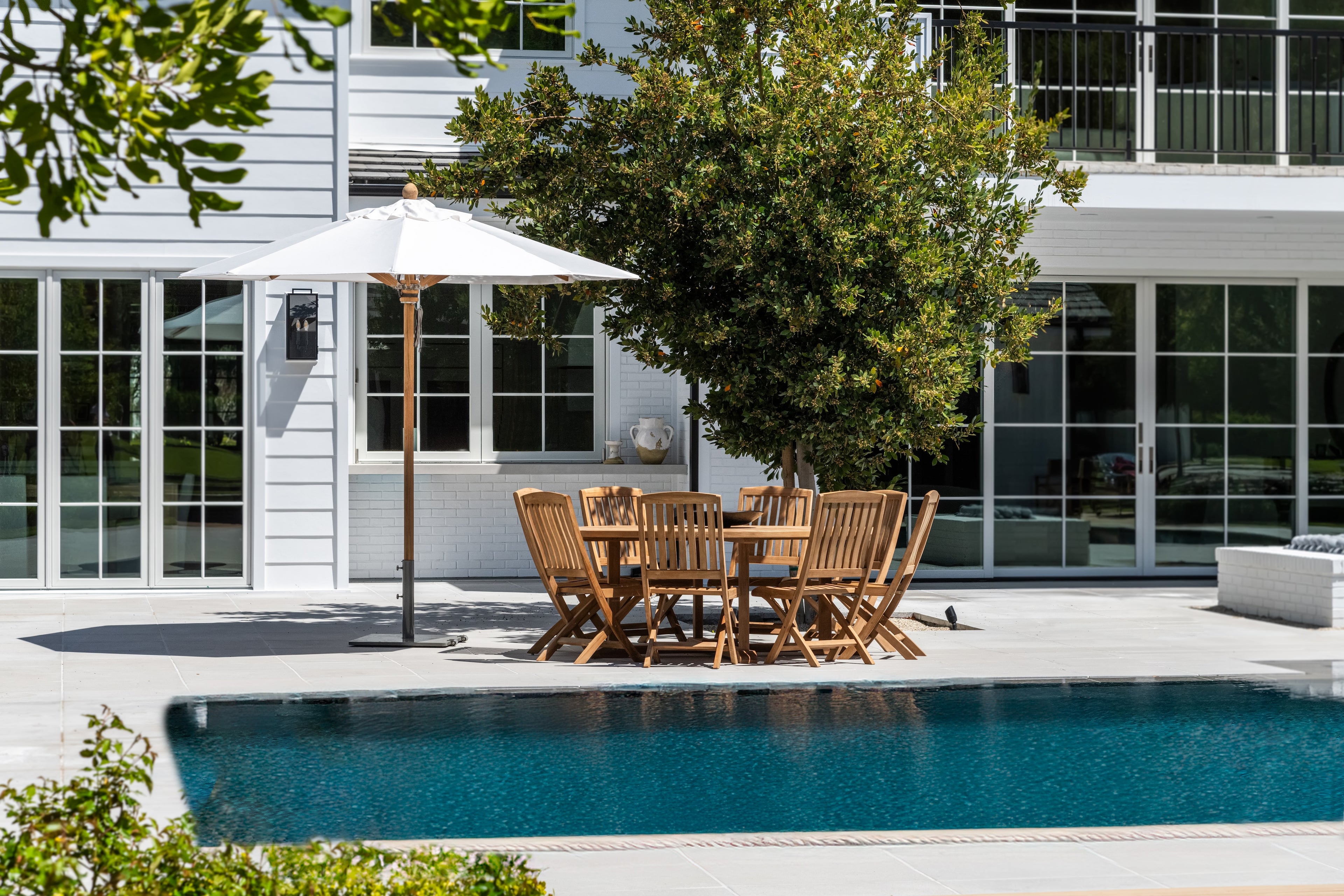 A wooden patio table with chairs sits under a white umbrella beside a swimming pool, surrounded by greenery and a white building with large glass doors and windows.