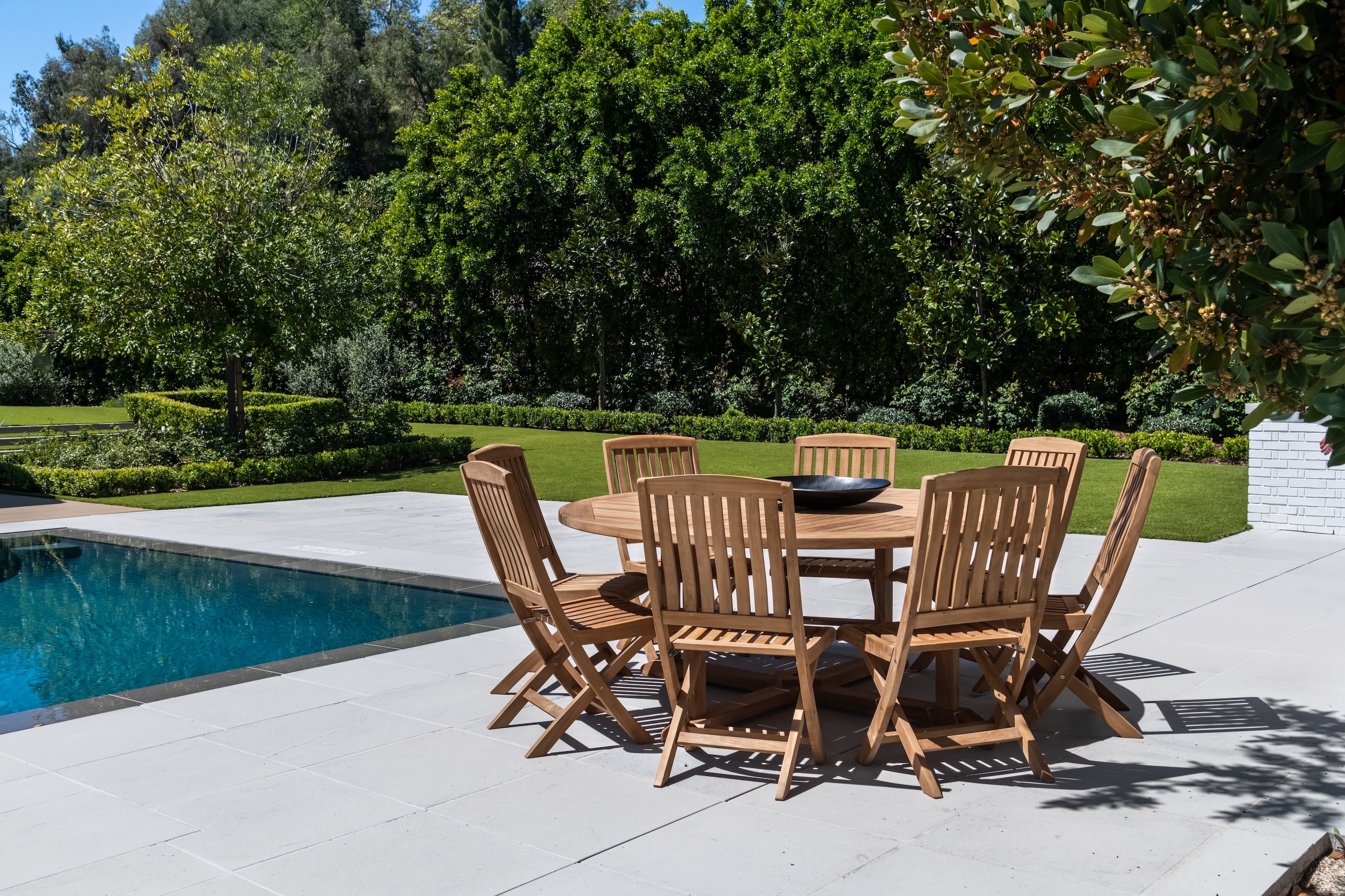 A wooden table with eight matching chairs is situated poolside on a sunny patio, surrounded by lush greenery and trees, creating a serene outdoor setting.