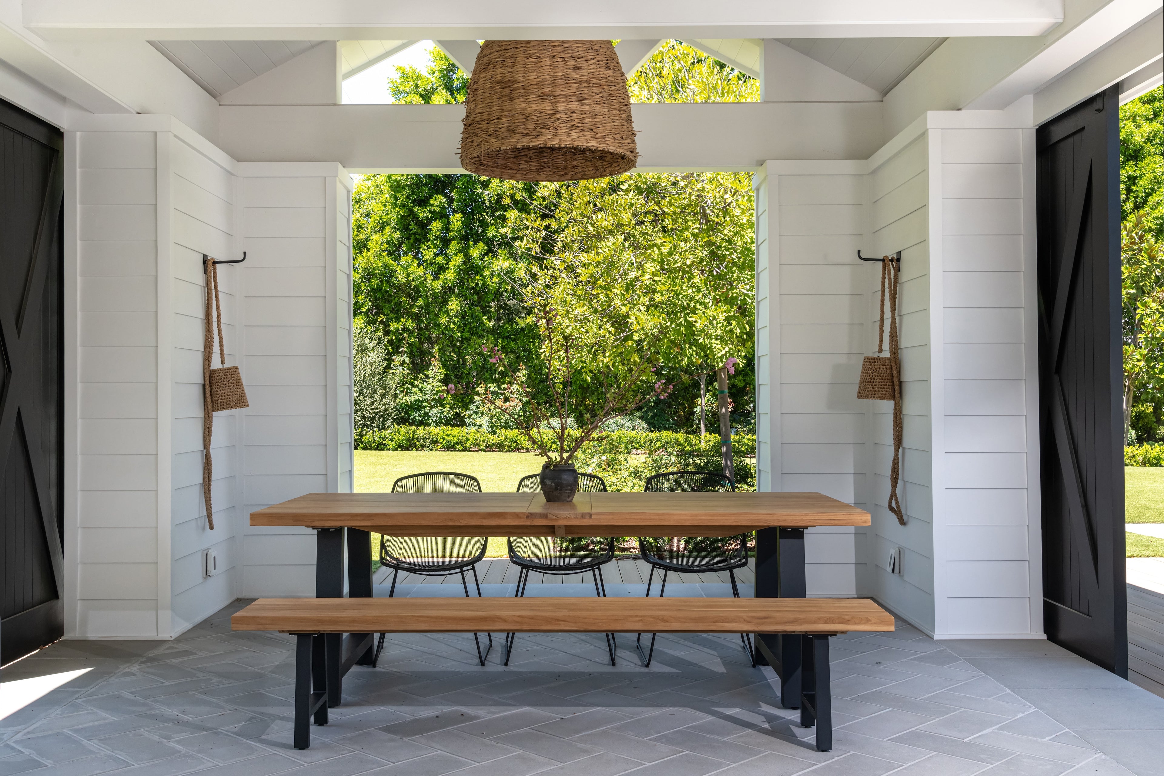 A wooden table with benches sits under a hanging wicker light fixture in a white, open-air room, overlooking a lush garden with trees and shrubs.