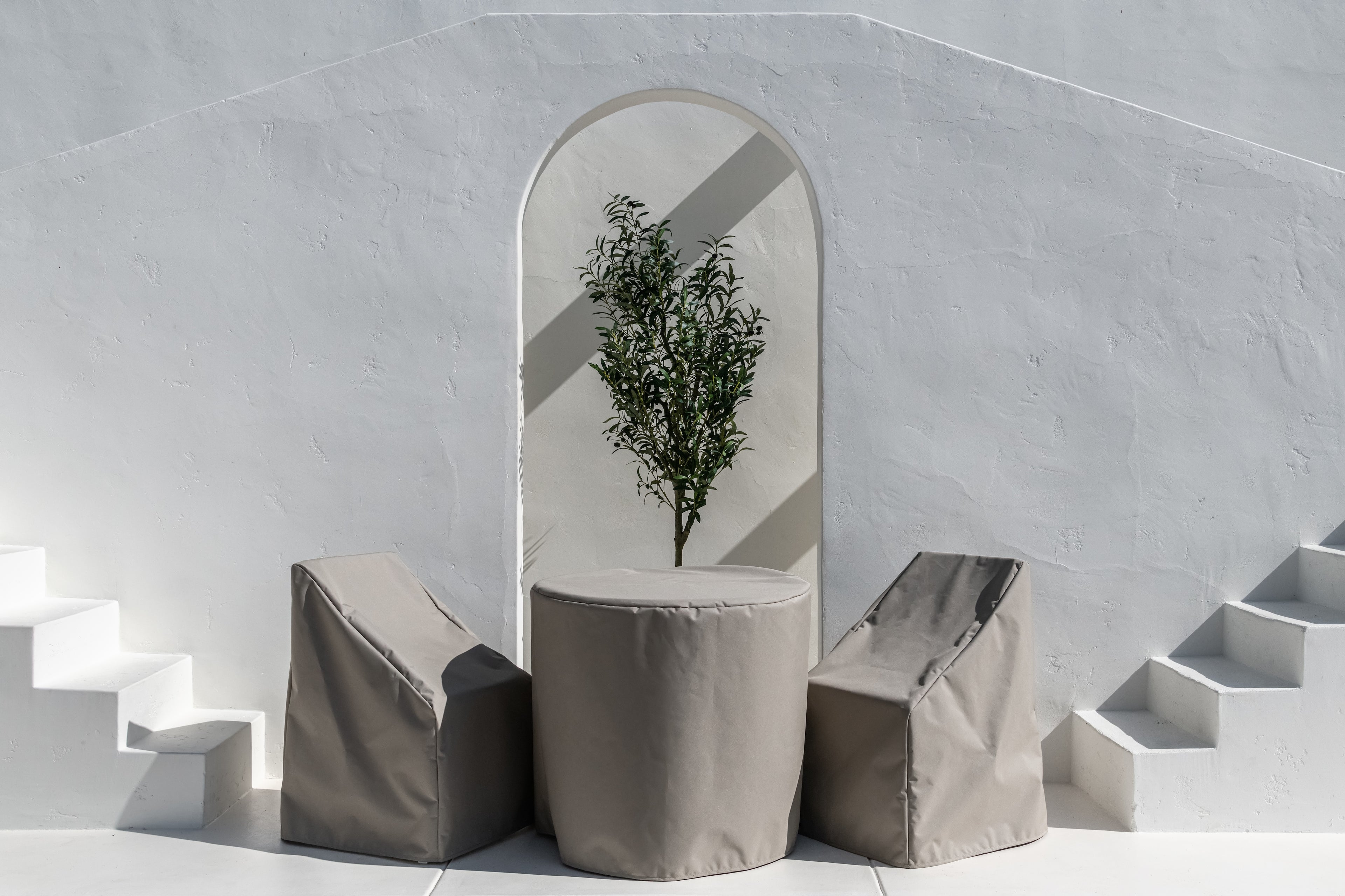 A round table and two angular chairs, covered with beige fabric, sit beneath an archway framing a small tree. They are positioned between white staircases against a textured wall.