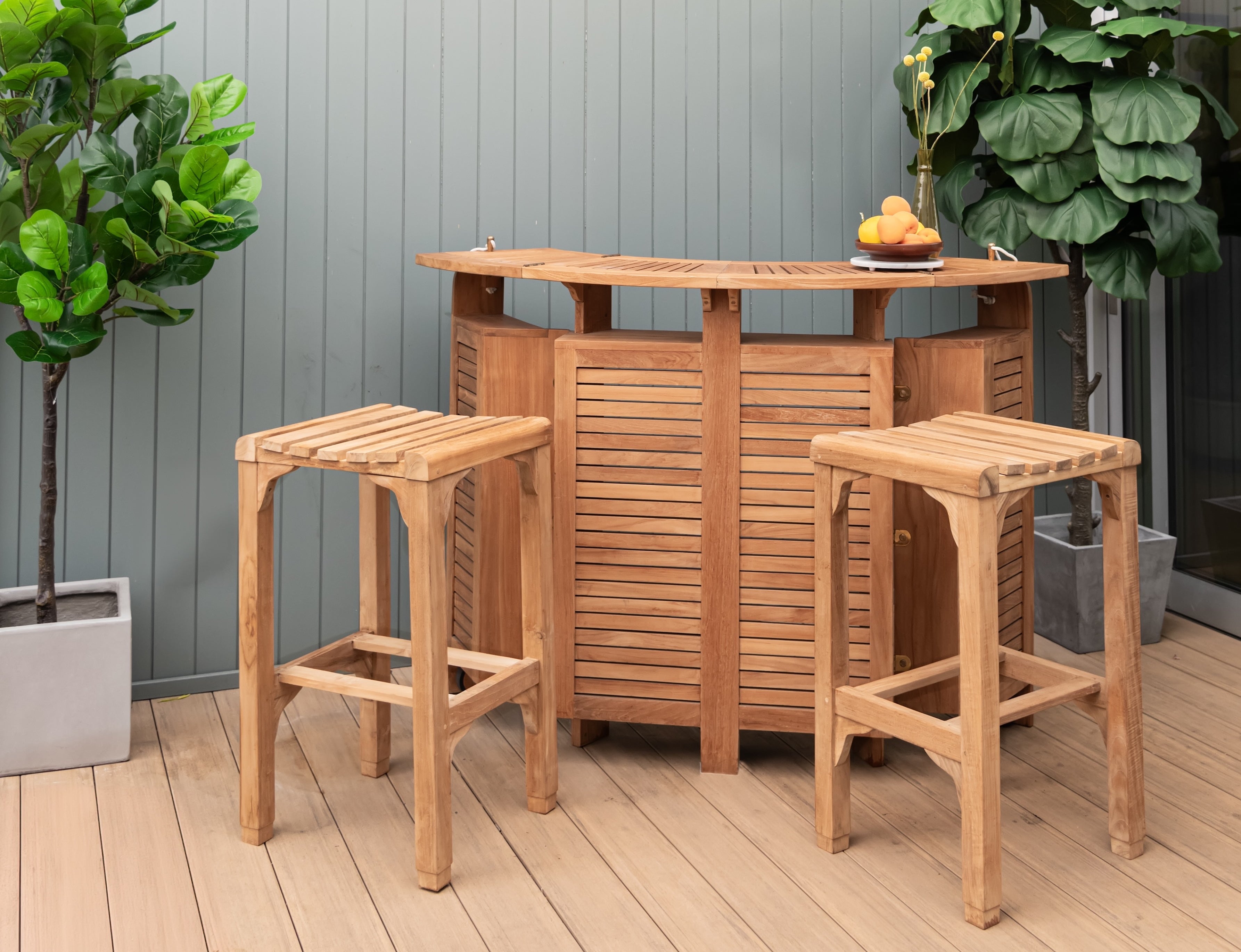 A wooden bar table is situated against a light green wall with two matching stools and plants in pots. A vase with flowers and fruit adds a decorative touch.