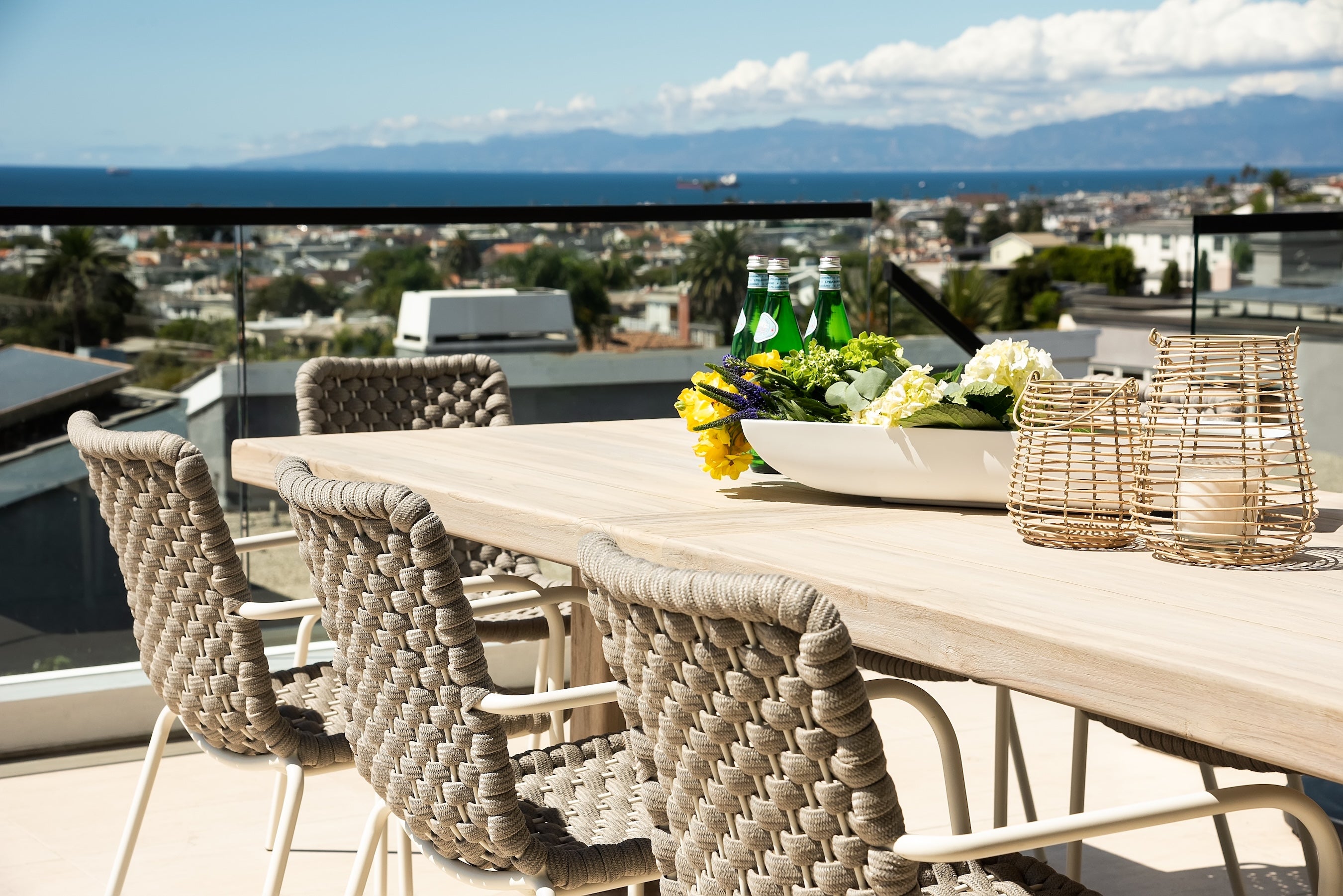 A wooden table holds flowers, glass bottles, and wicker baskets on a patio. Woven chairs surround it, overlooking a cityscape and ocean in the background.