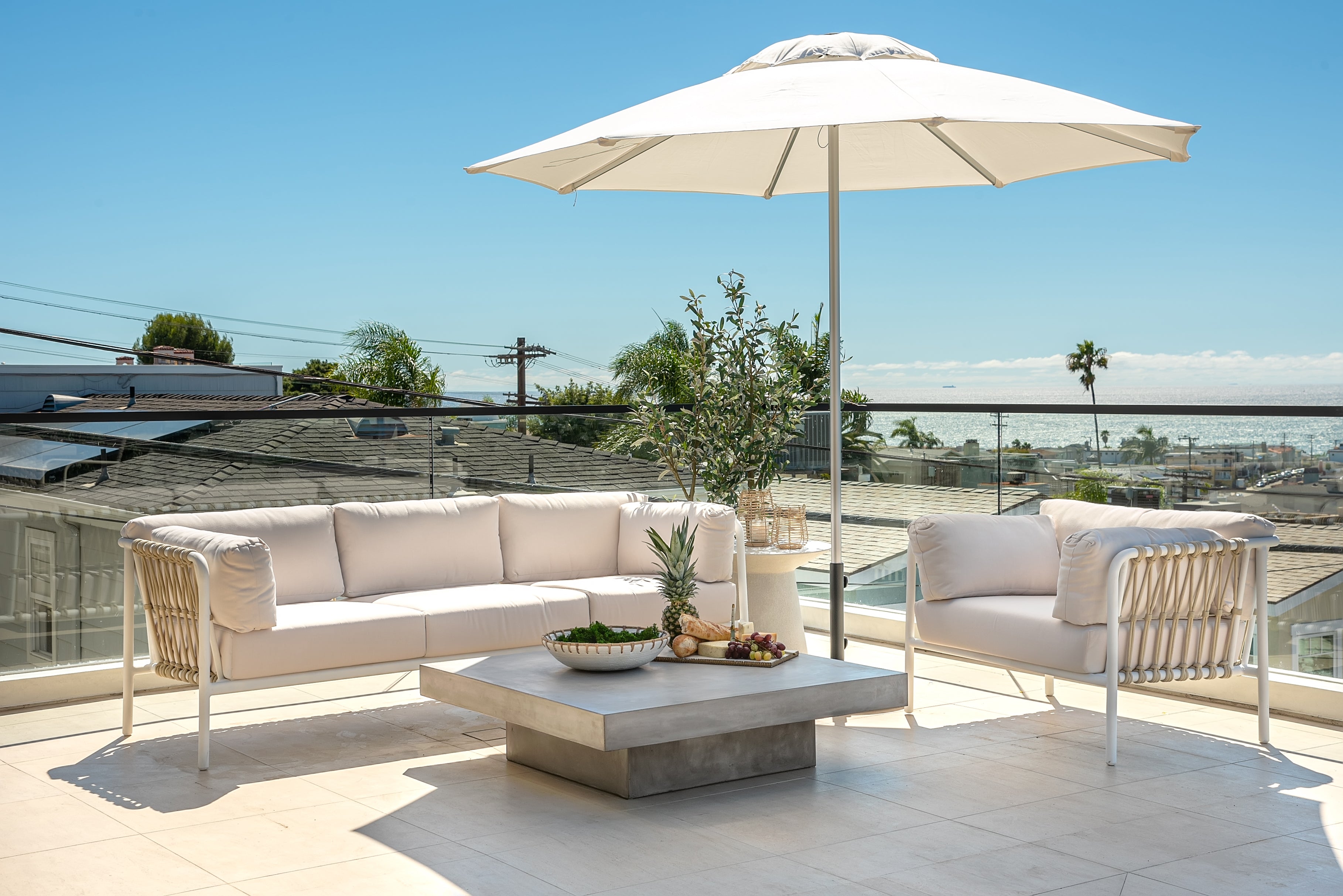 Outdoor patio with white cushioned seating, large umbrella, and square table featuring fruit. Overlooks rooftops with ocean in the distance, under a clear blue sky.