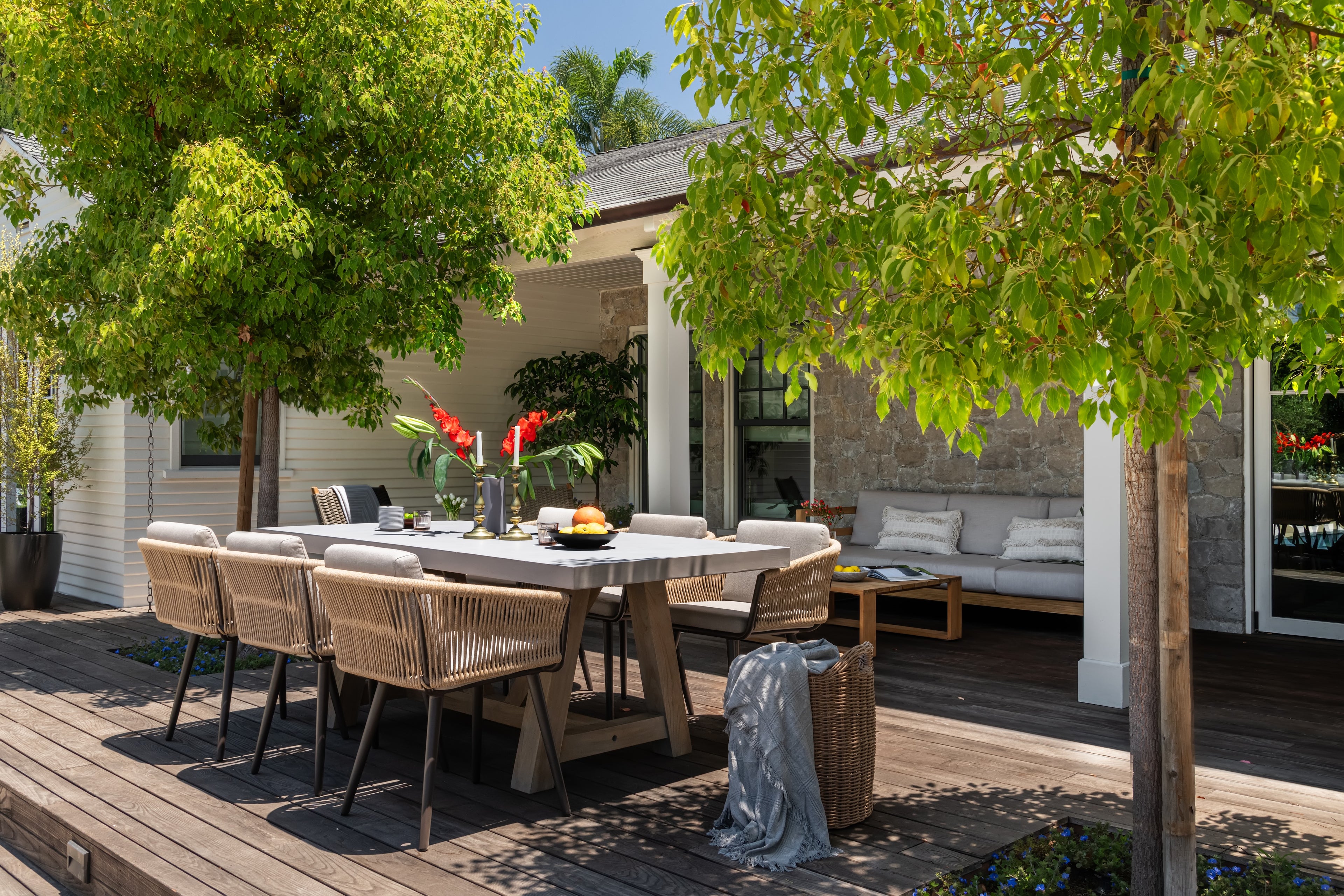 A wooden patio hosts a table set with red flowers and fruit. Surrounding chairs are shaded by lush trees. Nearby, a cozy sofa rests against a stone wall.