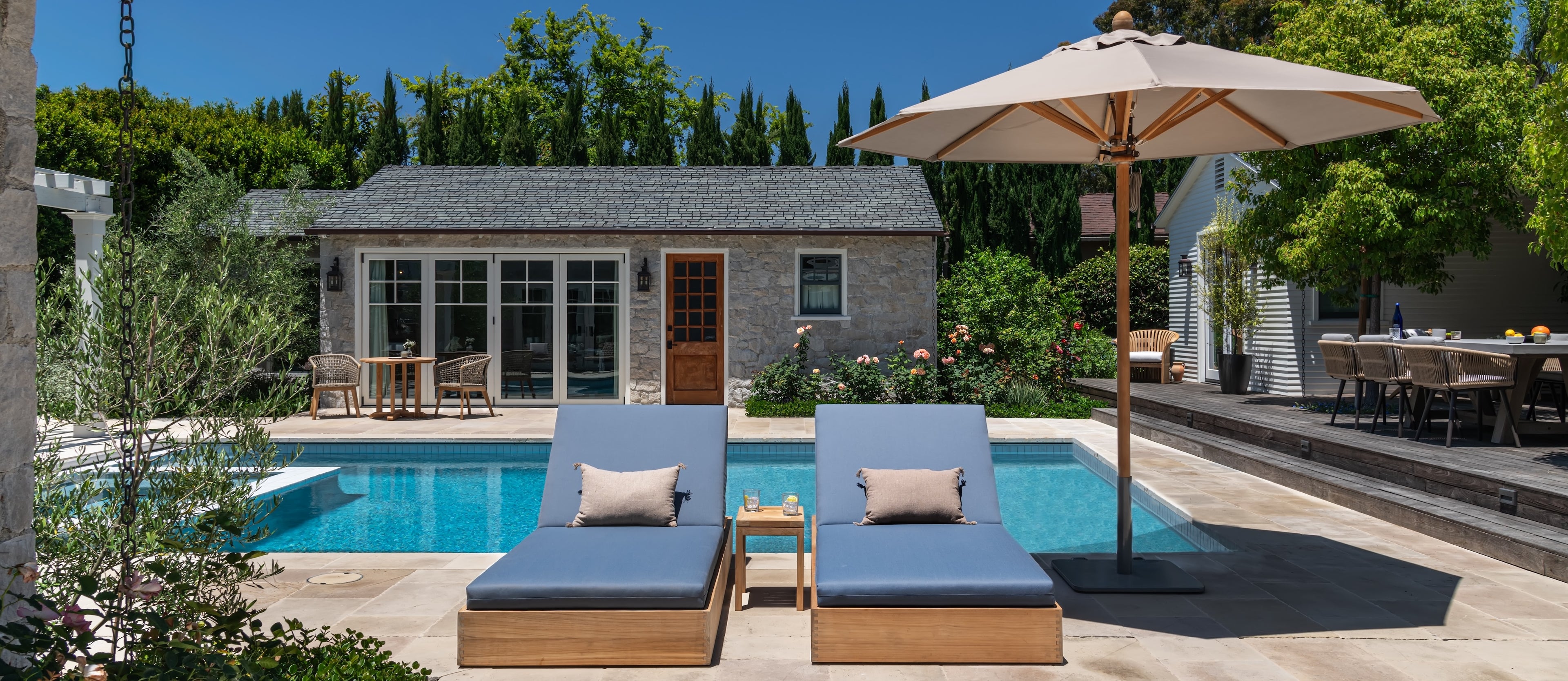 Two wooden lounge chairs sit by a pool, under an umbrella. A stone house with large windows and door is in the background, surrounded by plants and outdoor furniture.