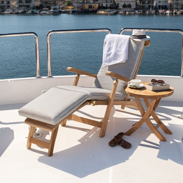 A wooden deck chair with a towel and hat rests beside a small table holding sunglasses, a cup, and a book, set