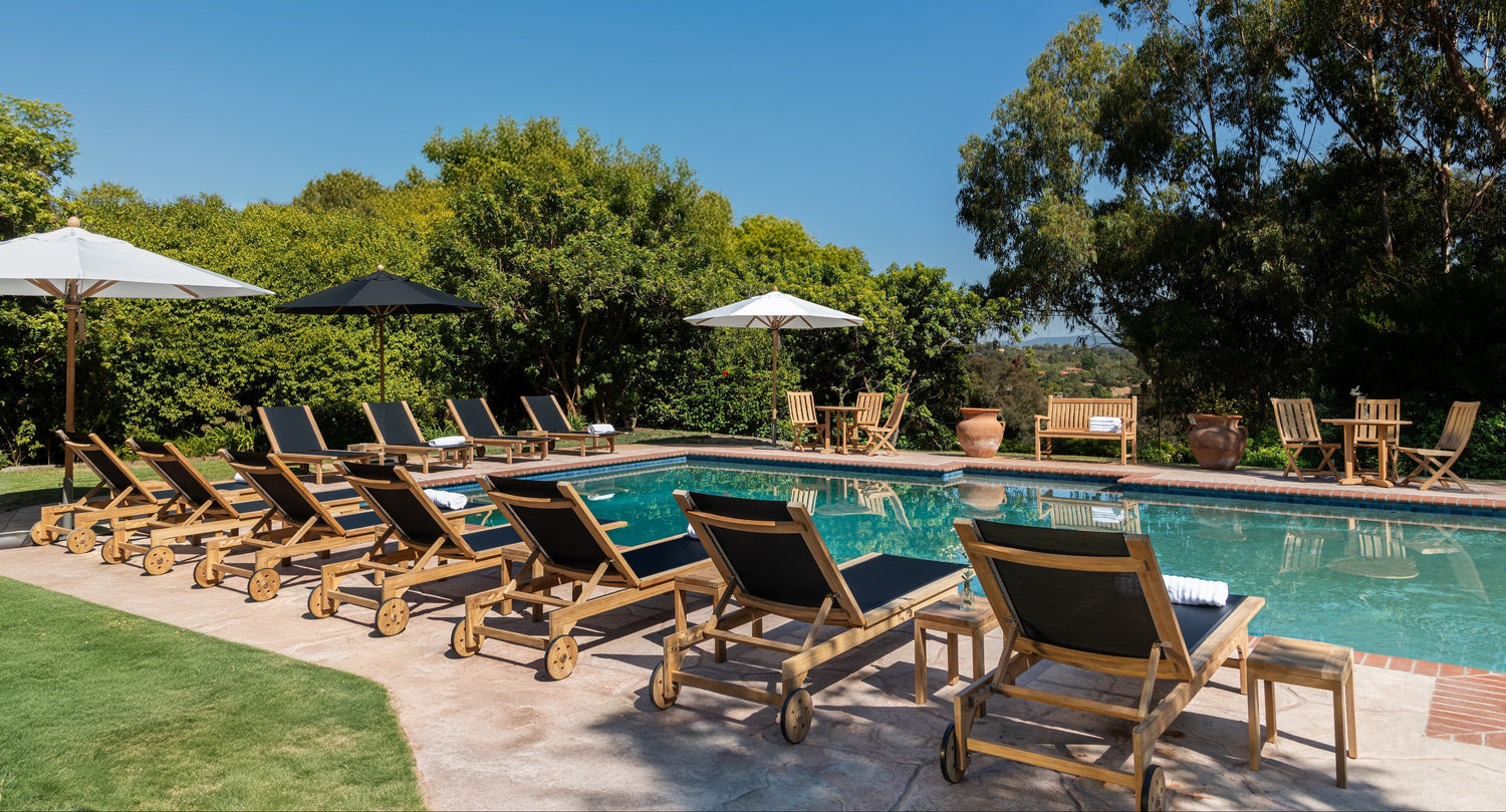 Wooden lounge chairs lined up along a pool; surrounded by umbrellas, tables, and greenery; under a clear blue sky.