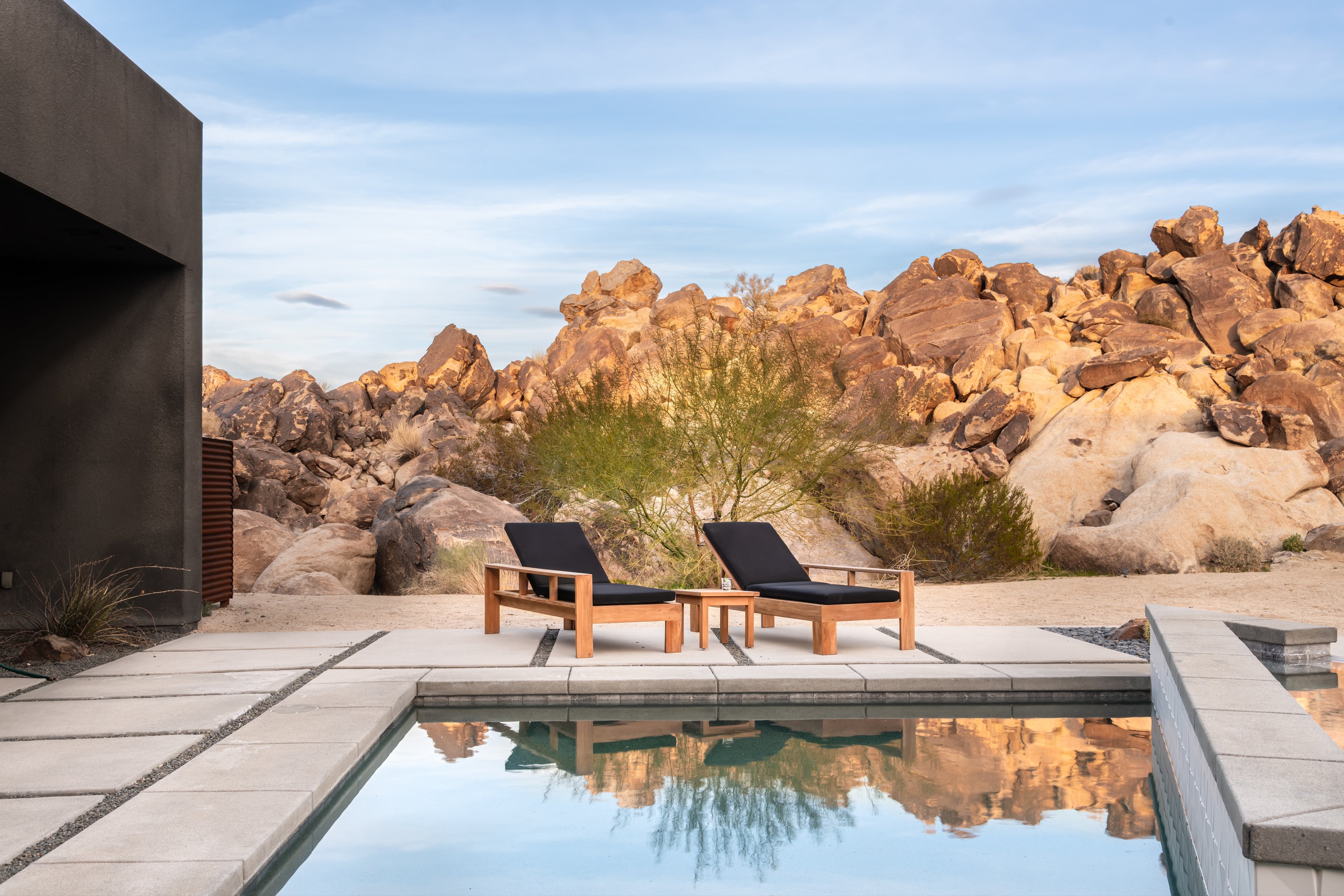 Poolside chairs sit empty beside a pool, with a small table in between. The setting features a rocky, desert landscape under a clear blue sky.