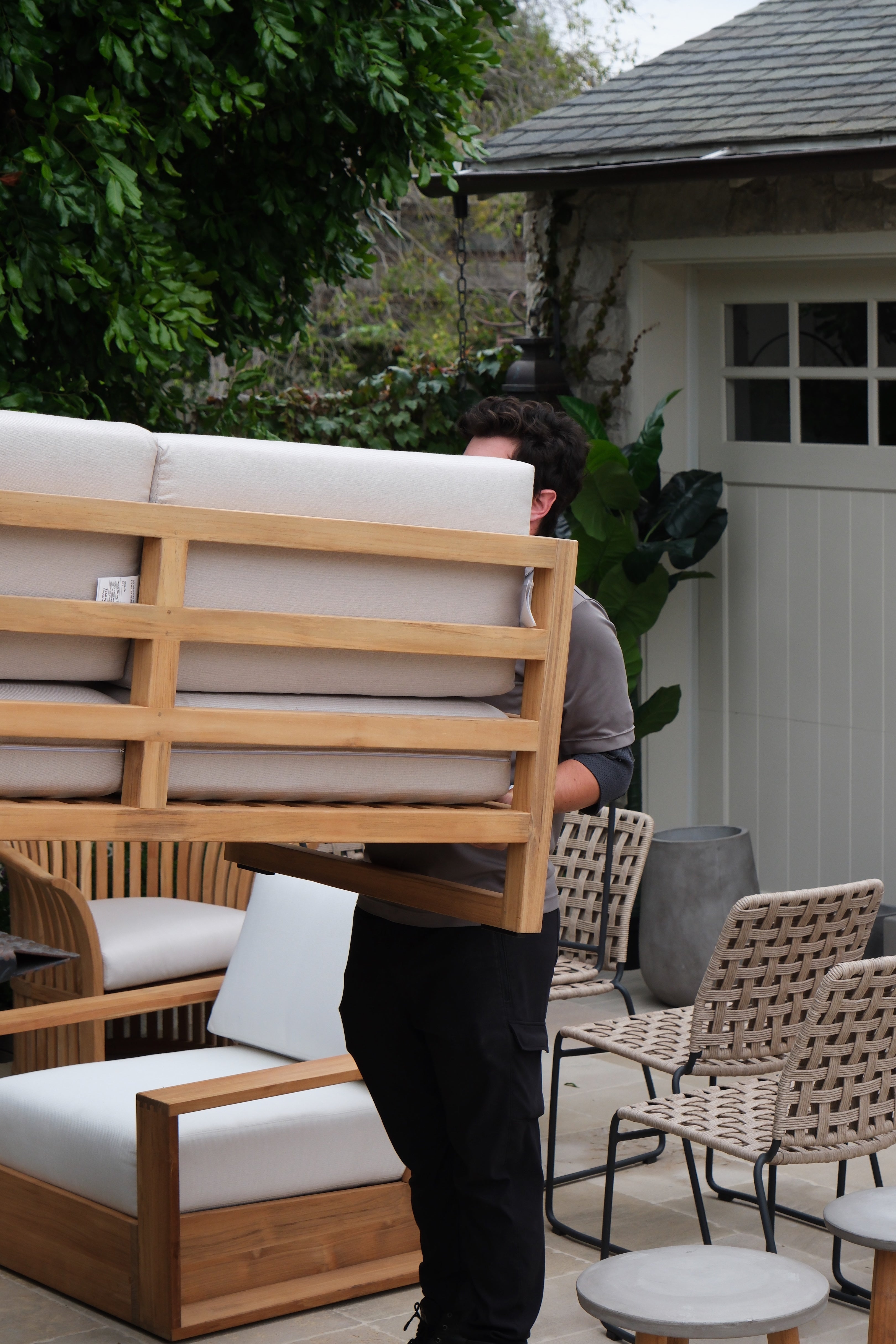 A person carries a wooden outdoor sofa with beige cushions. The setting includes patio furniture, a potted plant, and a house with a white door and greenery.