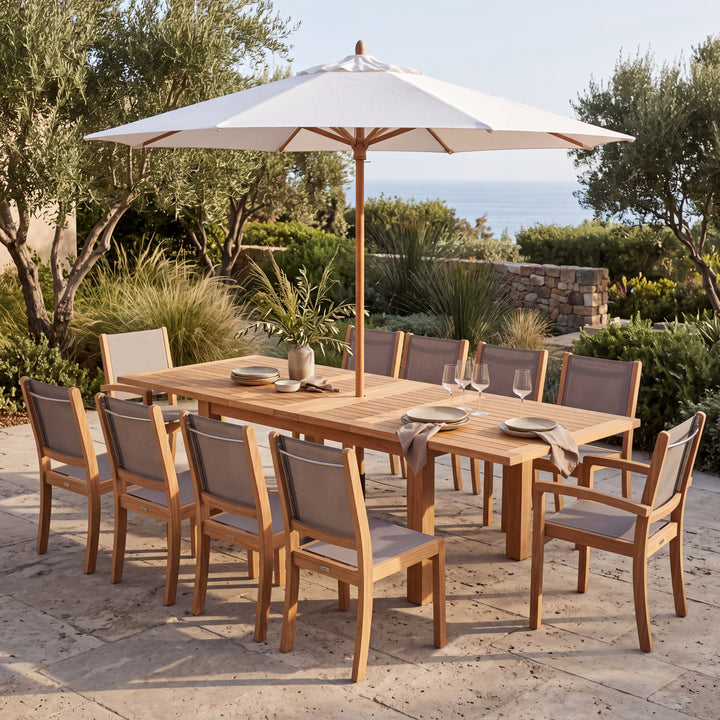 An outdoor dining set on a stone patio with a large white patio umbrella, featuring a rectangular wooden table and ten matching chairs with gray cushions, set against a garden backdrop with the ocean in the distance.