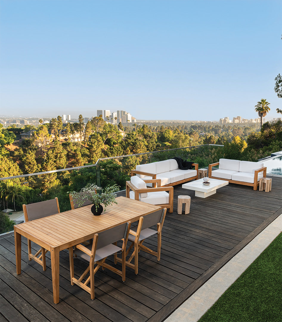 Wooden deck furniture set with chairs and a table is positioned on a terrace overlooking lush greenery and a distant city skyline under a clear blue sky.