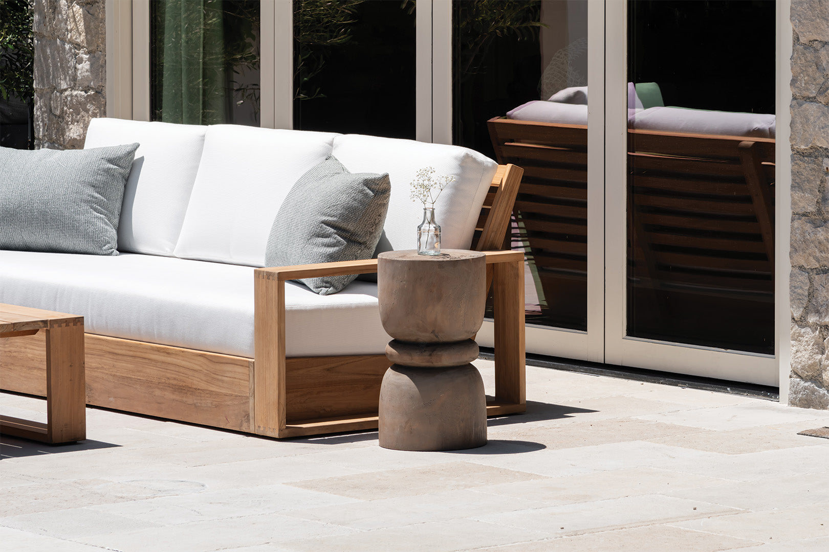 A wooden outdoor sofa with white cushions rests nearby a stone wall. A small, round wooden side table holds a clear vase with white flowers. Glass doors reflect nearby furniture.