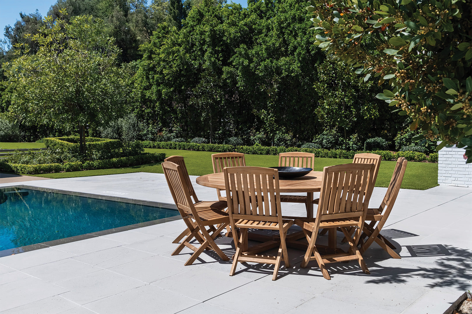 A wooden table surrounded by eight chairs sits on a patio beside a swimming pool, with a lush garden and tall trees in the background.