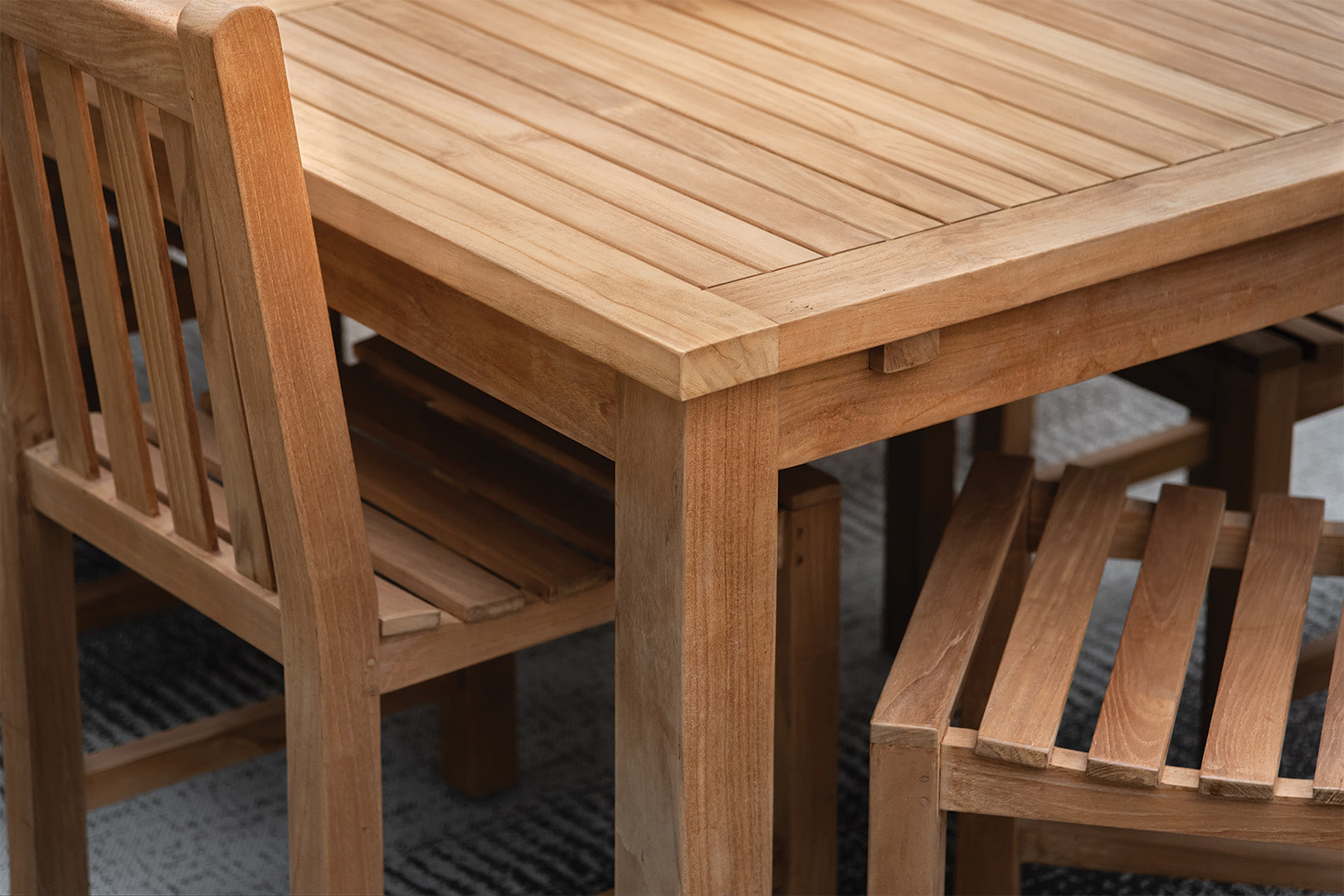 A wooden table with a slatted design is surrounded by matching chairs on a patterned rug. The furniture appears robust, suitable for an outdoor or indoor setting.