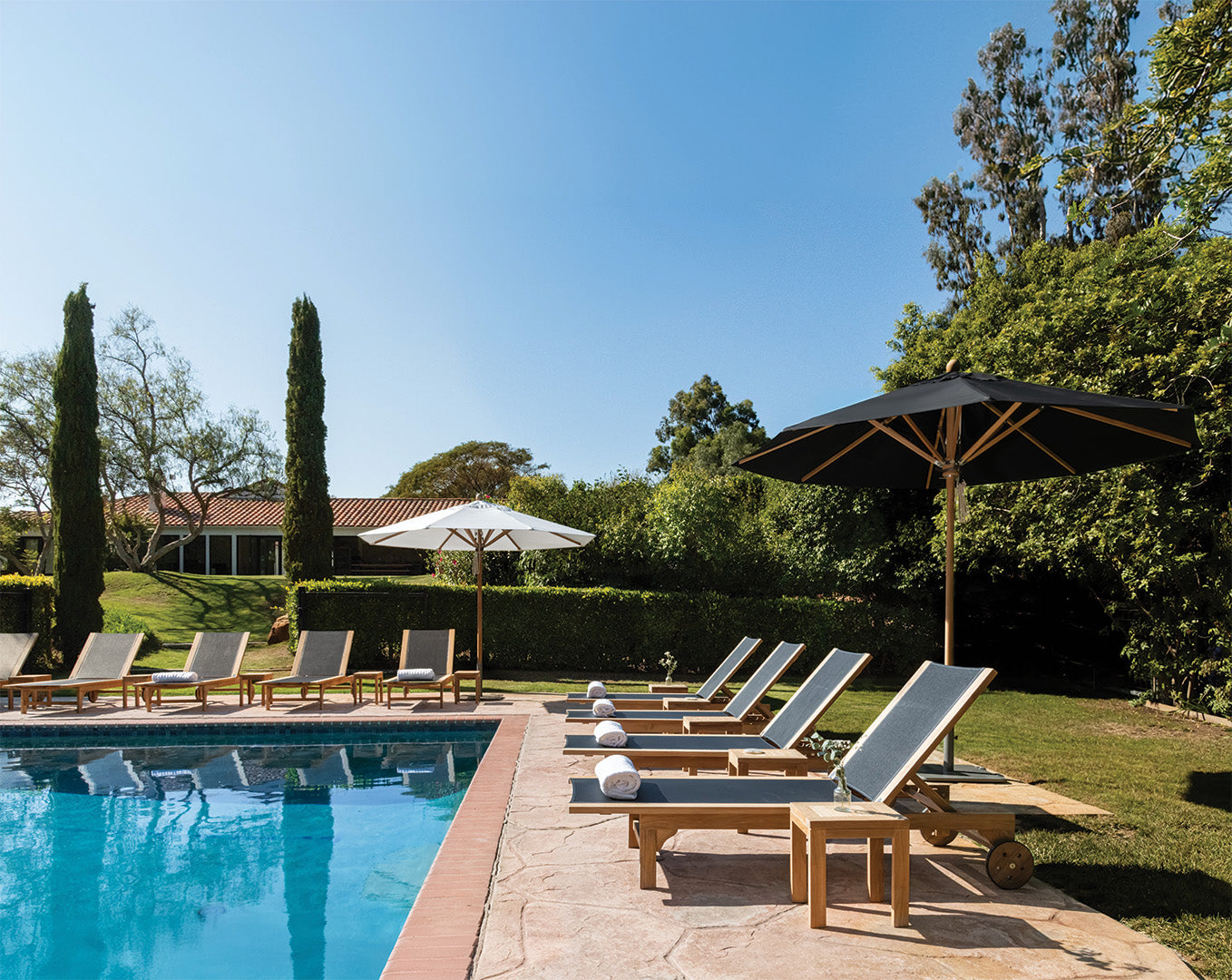 Lounge chairs are neatly arranged beside a calm swimming pool, under large umbrellas. The surroundings feature lush greenery and a distant house with a tiled roof, under a clear blue sky.