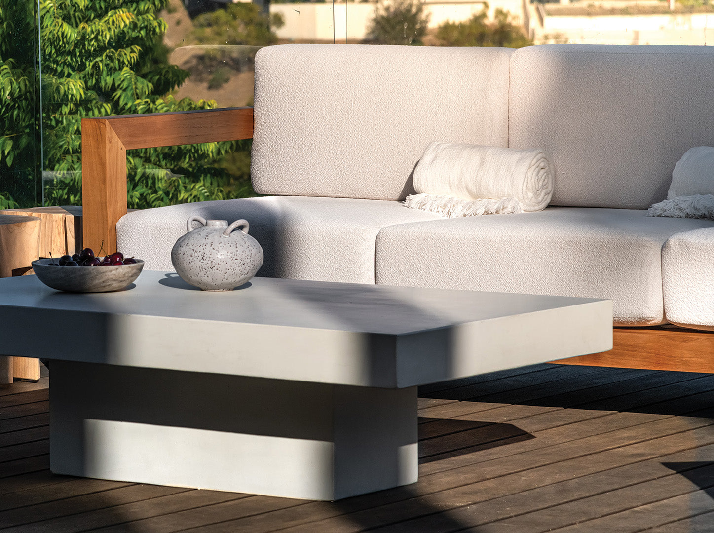 A white cushioned sofa with wooden arms sits on a wooden deck, with a modern, low rectangular table holding a bowl of berries and a speckled ceramic vase beside it, surrounded by greenery.