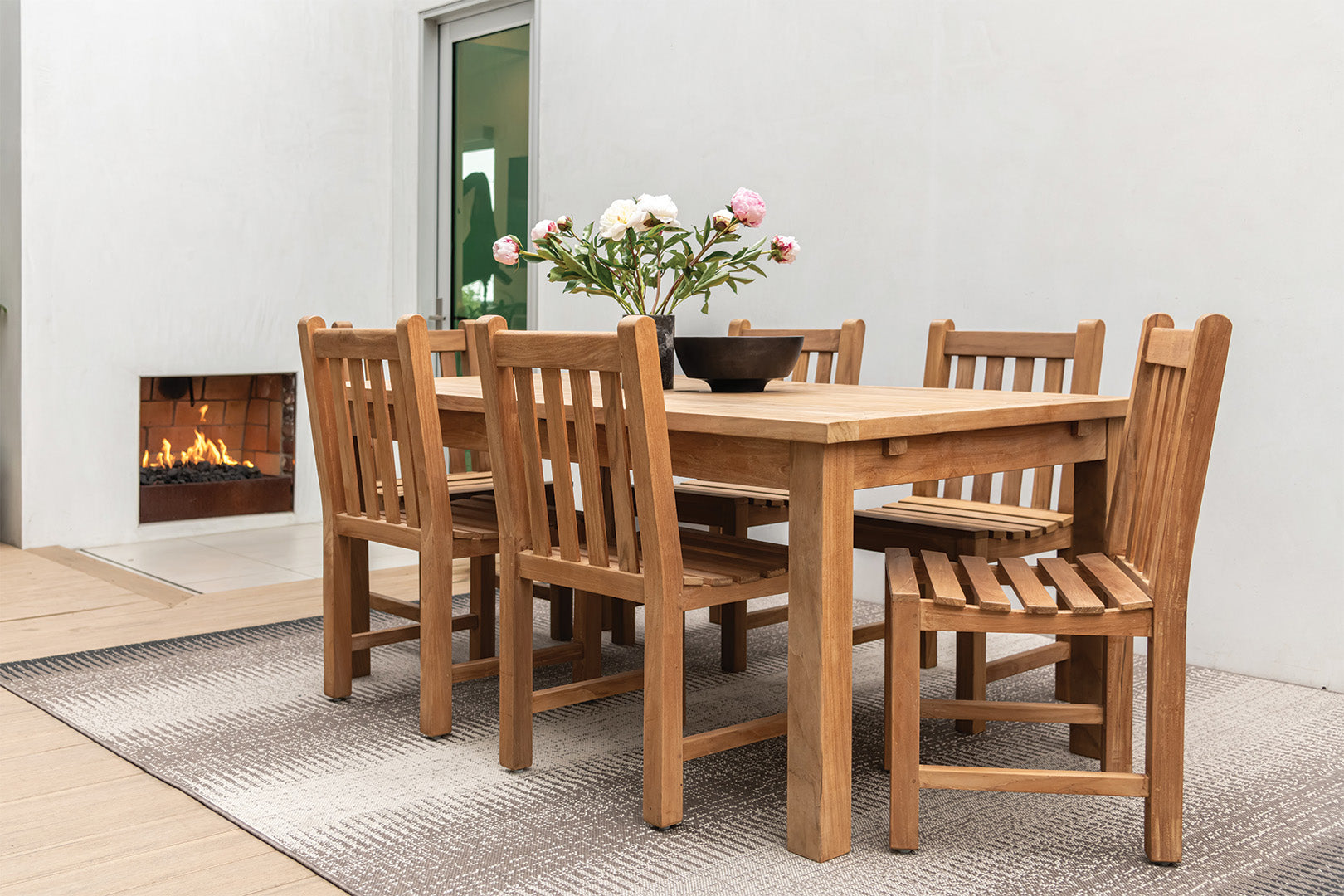 A wooden dining table with chairs displays a vase of flowers outdoors, set on a patterned rug beside a lit fireplace against a white wall.