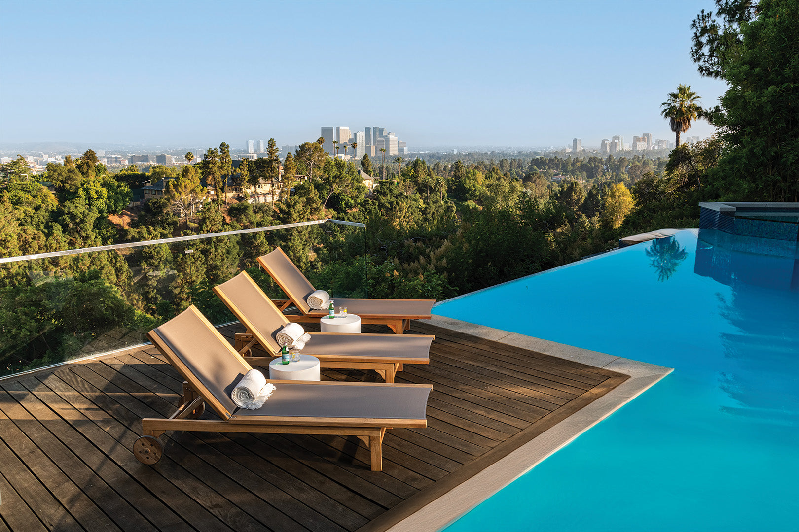 Three lounge chairs with rolled towels and drinks rest on a wooden deck beside an infinity pool, overlooking a lush green landscape and a distant city skyline under a clear blue sky.