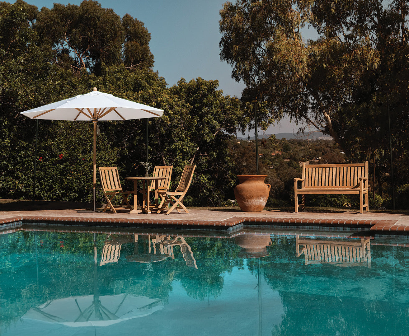 A wooden table with chairs and a white umbrella sits by a clear pool. A bench and large pot are nearby, surrounded by lush green trees under a clear sky.