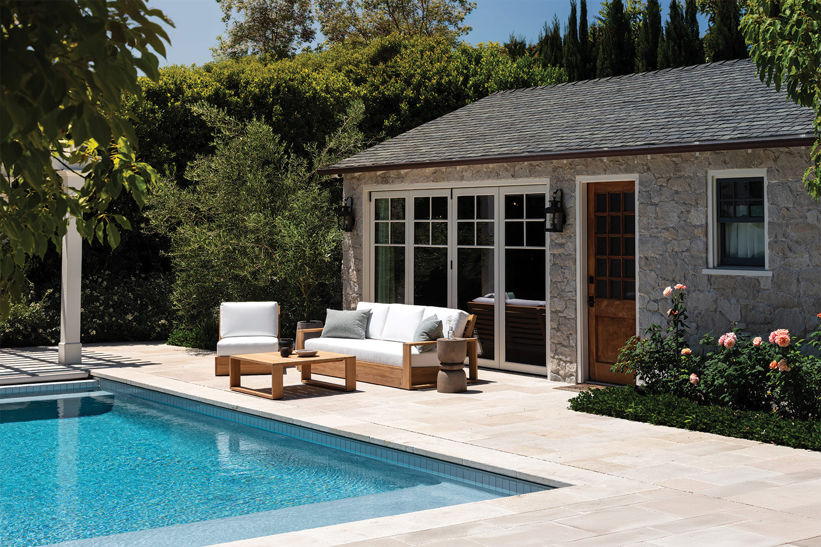 Wooden patio furniture sits by a swimming pool, featuring a couch and table. The setting includes a stone house with glass doors, surrounded by lush greenery and flowering plants.