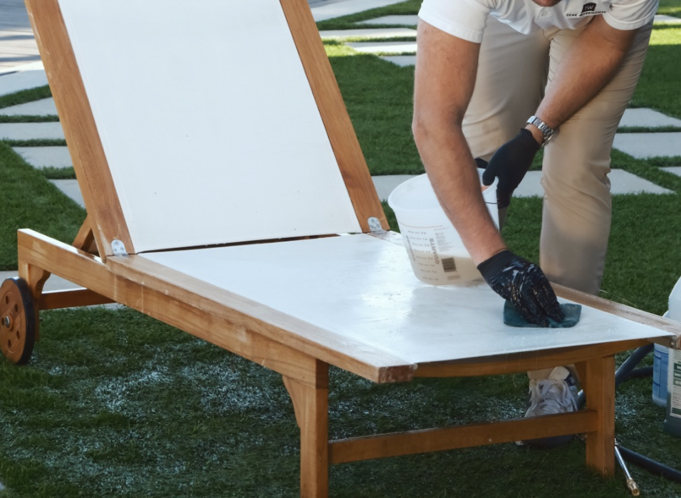 A person refinishes a wooden lounge chair, using a sponge, in a garden setting with grass and stepping stones, alongside a bucket marked with volume measurements.