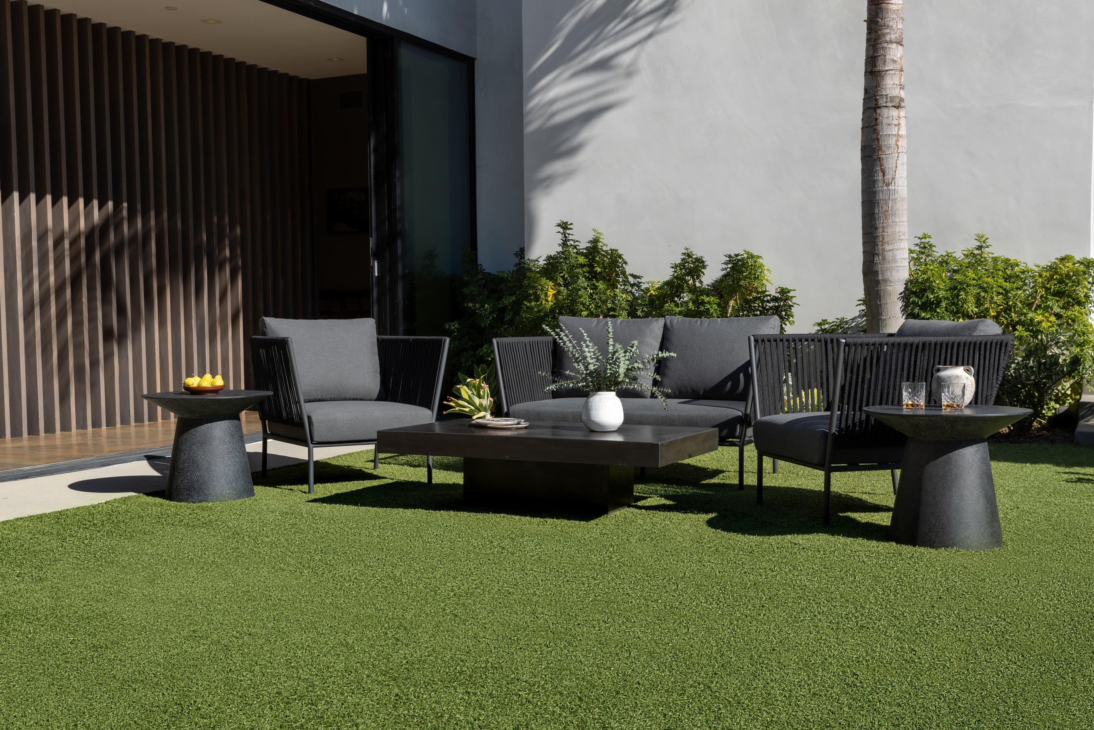 A patio set, featuring a black table with gray armchairs and a sofa, is placed on green artificial grass. A potted plant decorates the table, framed by a building and greenery.