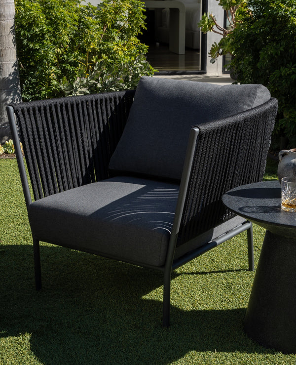 A black cushioned chair sits on a lawn beside a small round table holding a ceramic vase and two glasses, surr