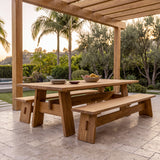 A large wooden outdoor dining table with two matching benches under a pergola, set on a stone patio with palm trees and greenery in the background at sunset.