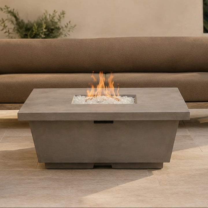 A rectangular concrete outdoor fire pit table with a burning flame and white stones inside, positioned in front of a modern tan outdoor sofa on a light stone patio.