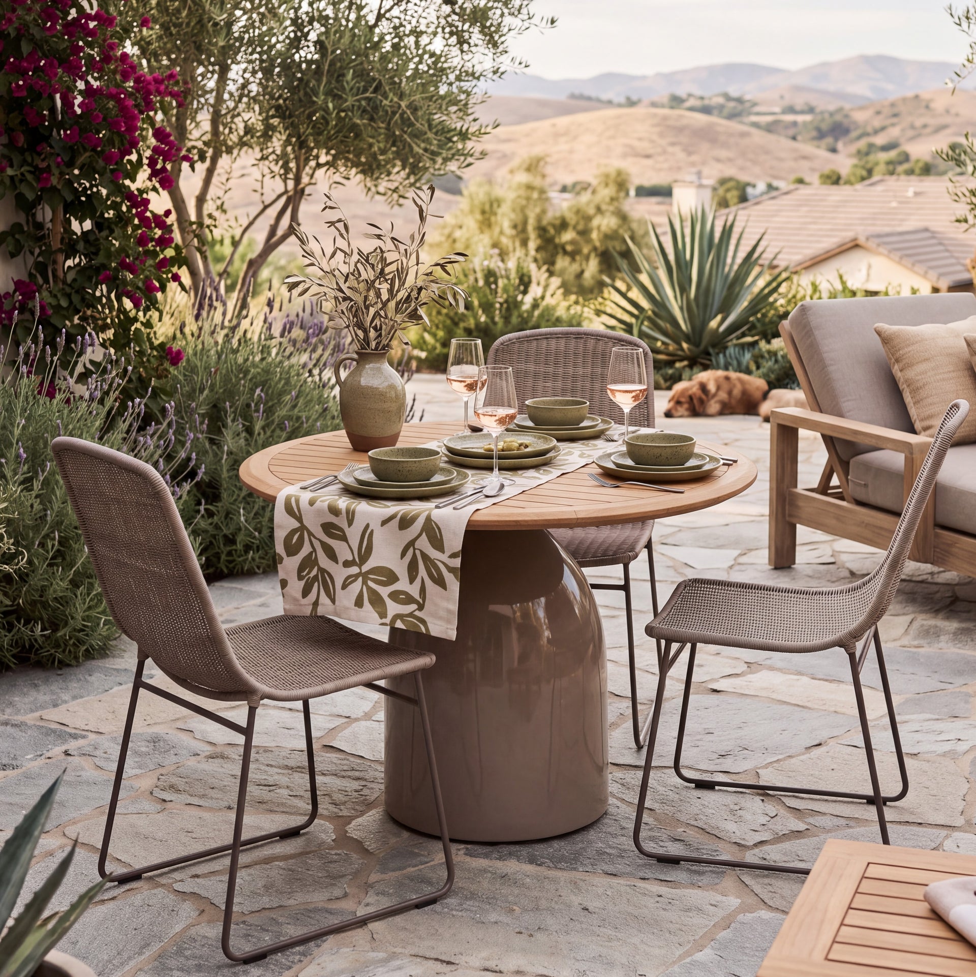 Outdoor patio dining setup with a round wooden table on a thick round pedestal base, surrounded by woven wicker chairs. The table is set with green bowls, plates, and a patterned runner.