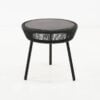 A round black table with a smooth top and a woven design on the sides stands on three slender legs in a minimalistic white space.